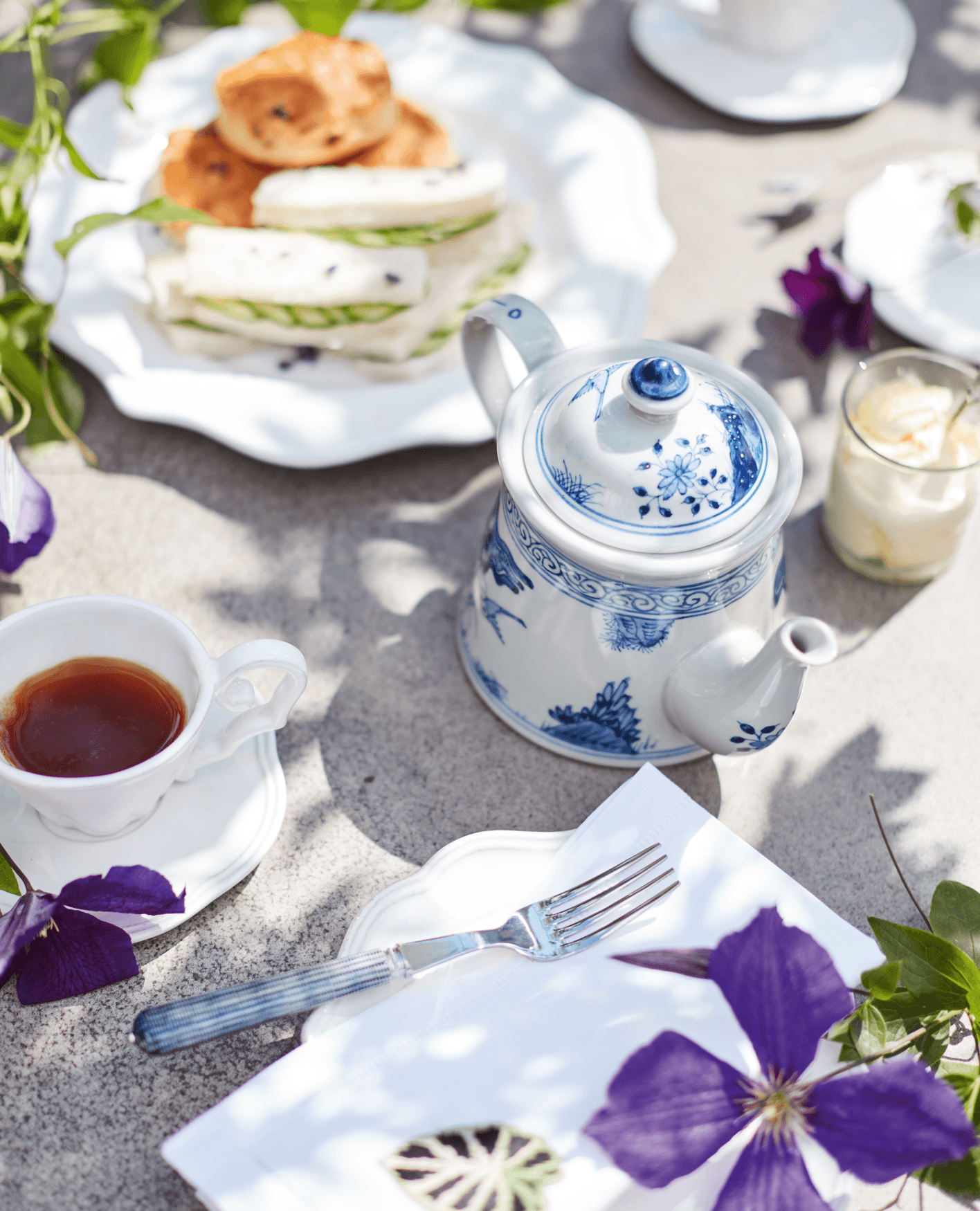An elegant afternoon tea, with a blue-and-white teapot, and a white plate holding scones and cucumber sandwiches.