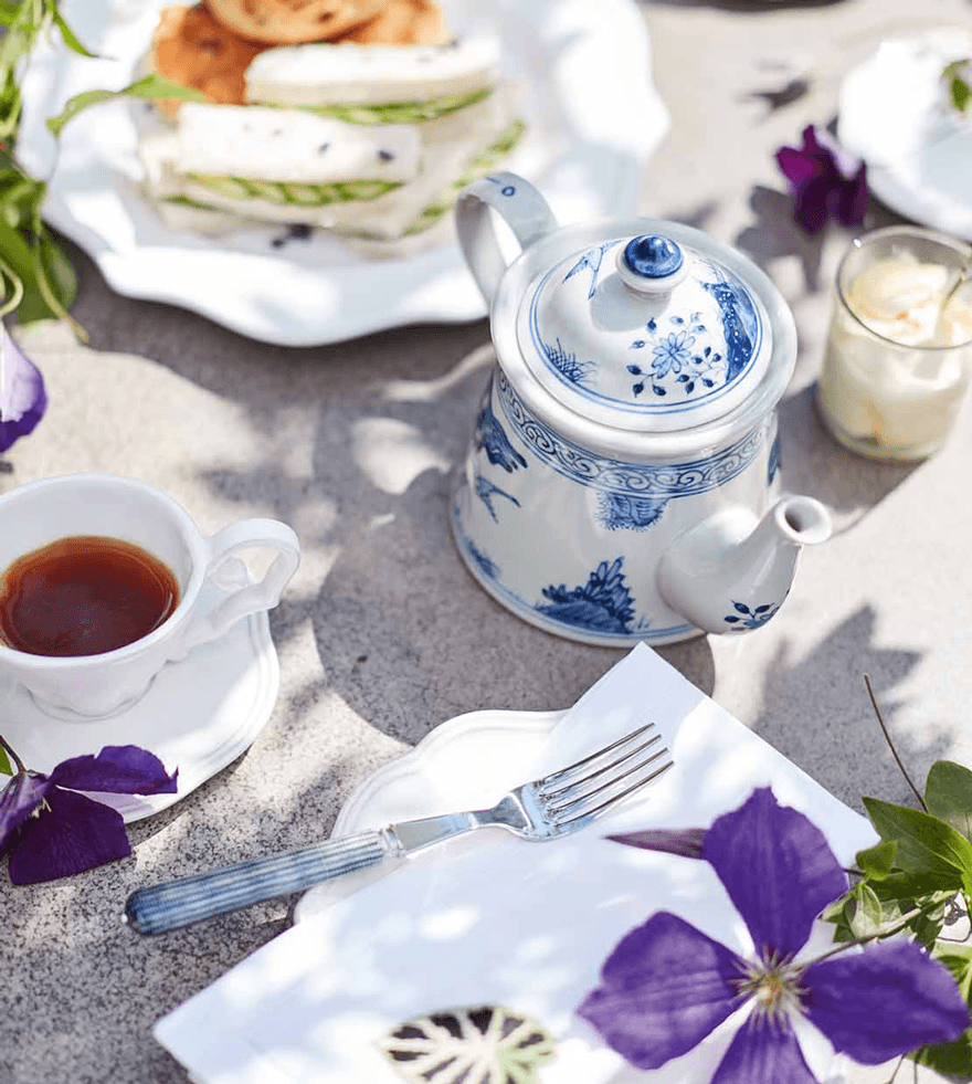 An elegant afternoon tea, with a blue-and-white teapot, and a white plate holding scones and cucumber sandwiches.