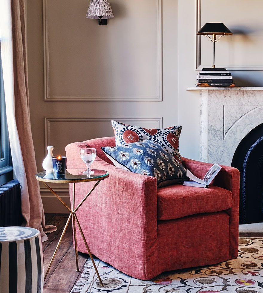 A corner of a living room, featuring a red armchair with two cushions on top, next to a glass side table, in front of a marble stone fire place.