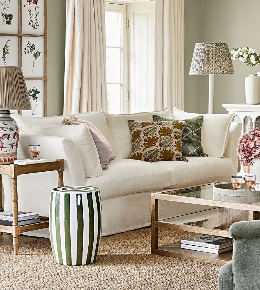 A corner of a living room, featuring a large white sofa, surrounded by side tables, a coffee table, lamps and cushions