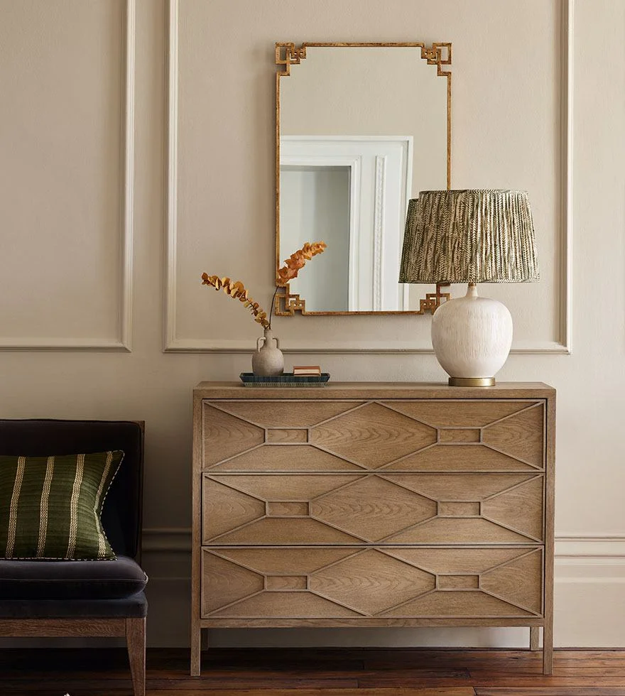 A rectangular mirror with metal gold frame up hanging on wall, in front of a chest drawers with a table lamp and a vase with flowers. 