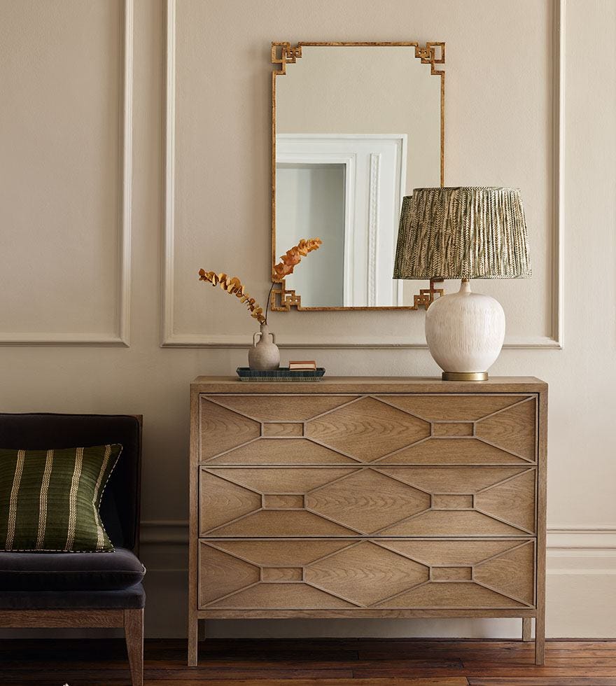 A gold mirror hangs on the wall, above a wooden dresser decorated with a white lamp.