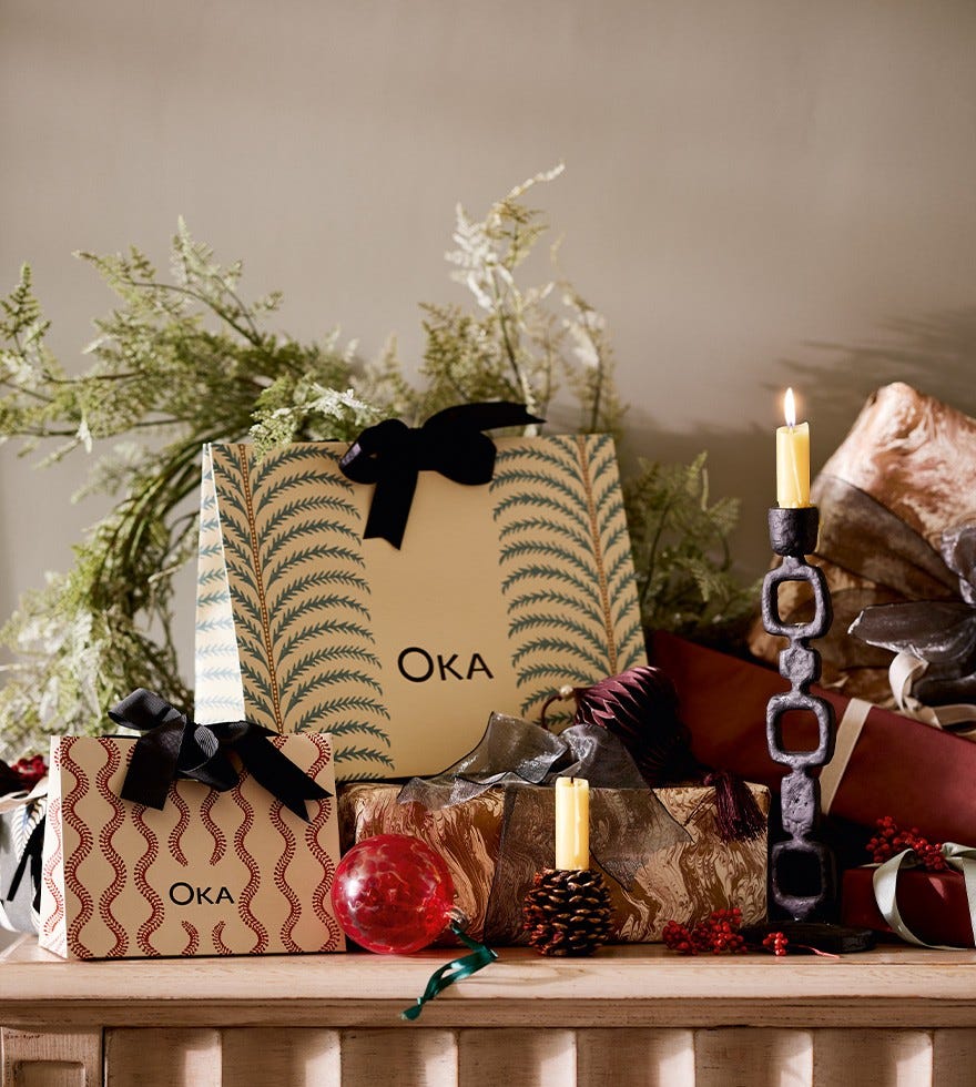 OKA gift bags are arranged on a table with foliage, candle holders, gifts and red patterned ornaments.