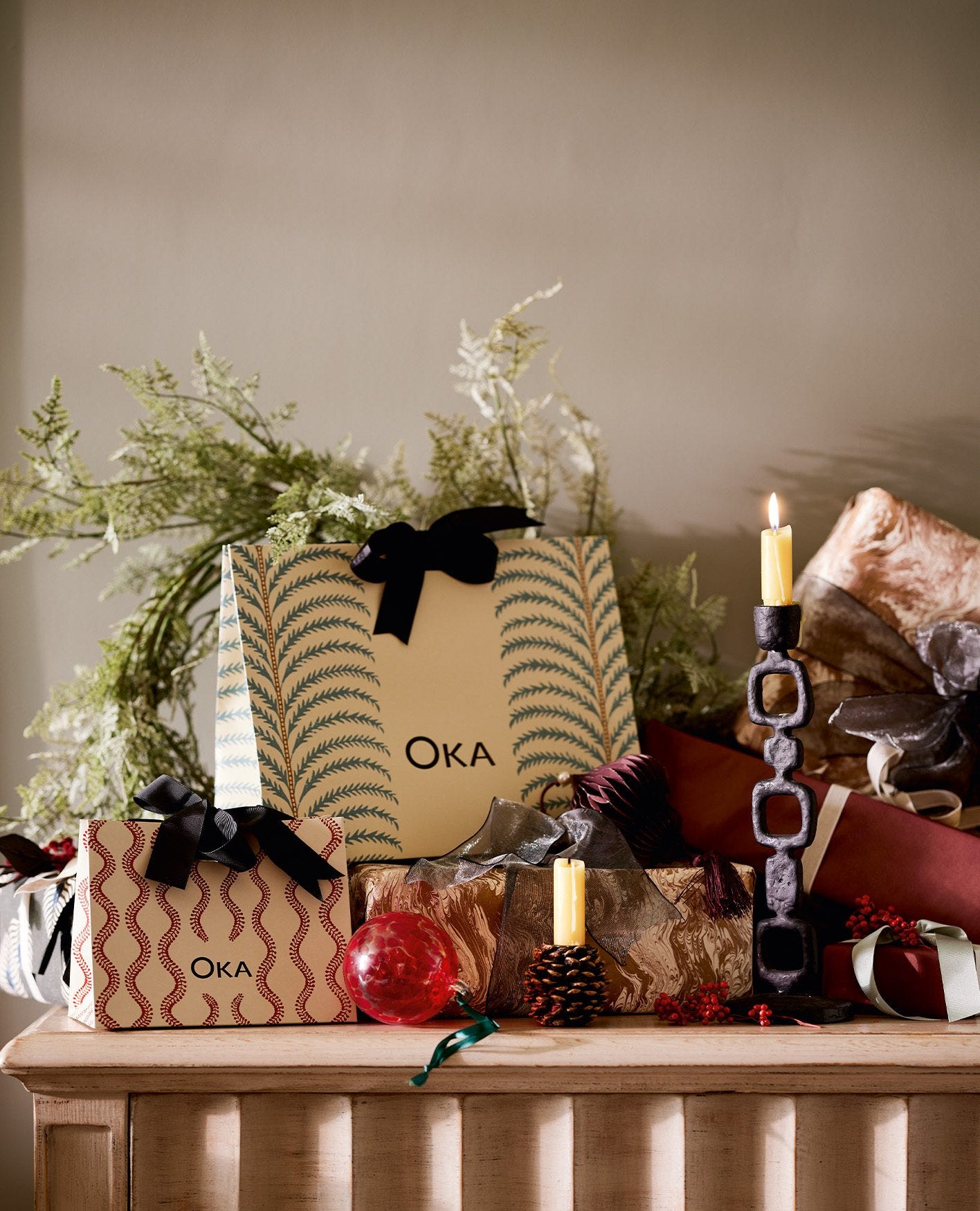 OKA gift bags are arranged on a table with foliage, candle holders, gifts and red patterned ornaments.