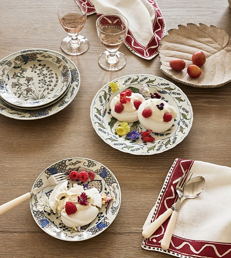 A wooden table is set with patterned plates holding meringues and berries. Red-and-white napkins, wine glasses and white flatware sit next to them.