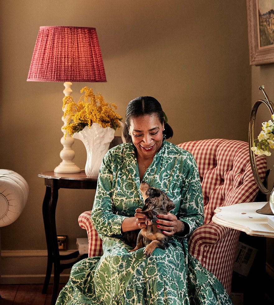 Paula Sutton sat on an armchair, beside a white side table decorated with a glass vase and flowers and a large table lamp behind her with a a large pleated lampshade.