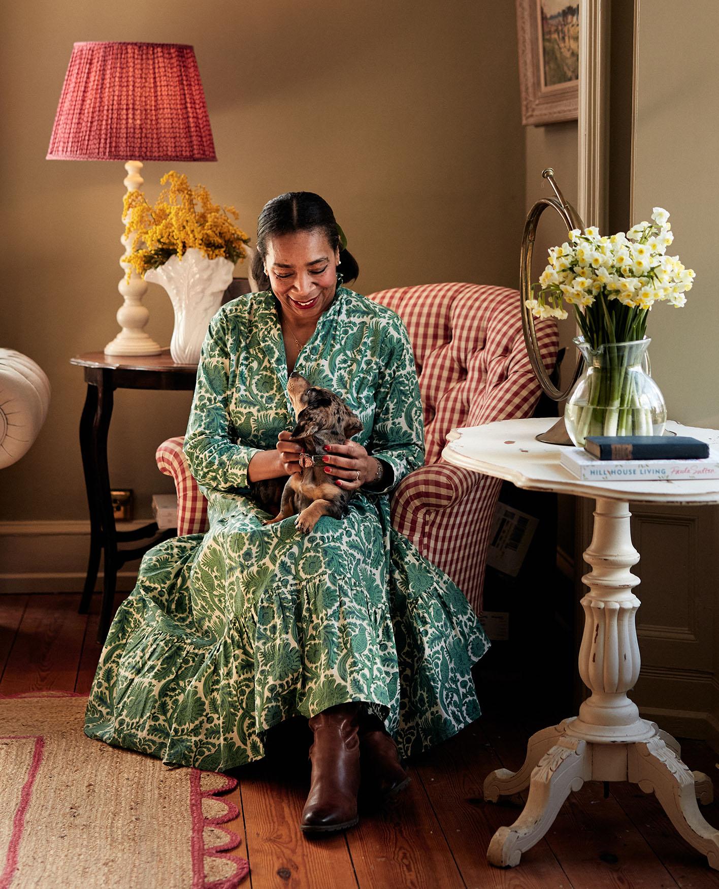 Paula Sutton sat on an armchair, beside a white side table decorated with a glass vase and flowers and a large table lamp behind her with a a large pleated lampshade.