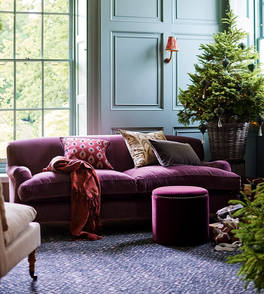 A bright magenta velvet sofa sits in a blue-walled room. A Christmas tree is seen in the background, and the sofa is decorated with cushions.