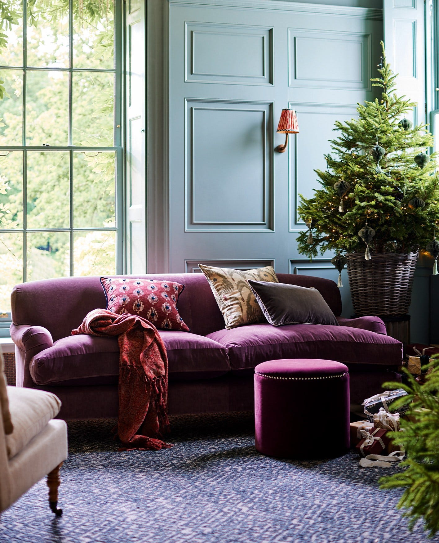 A bright magenta velvet sofa sits in a blue-walled room. A Christmas tree is seen in the background, and the sofa is decorated with cushions.