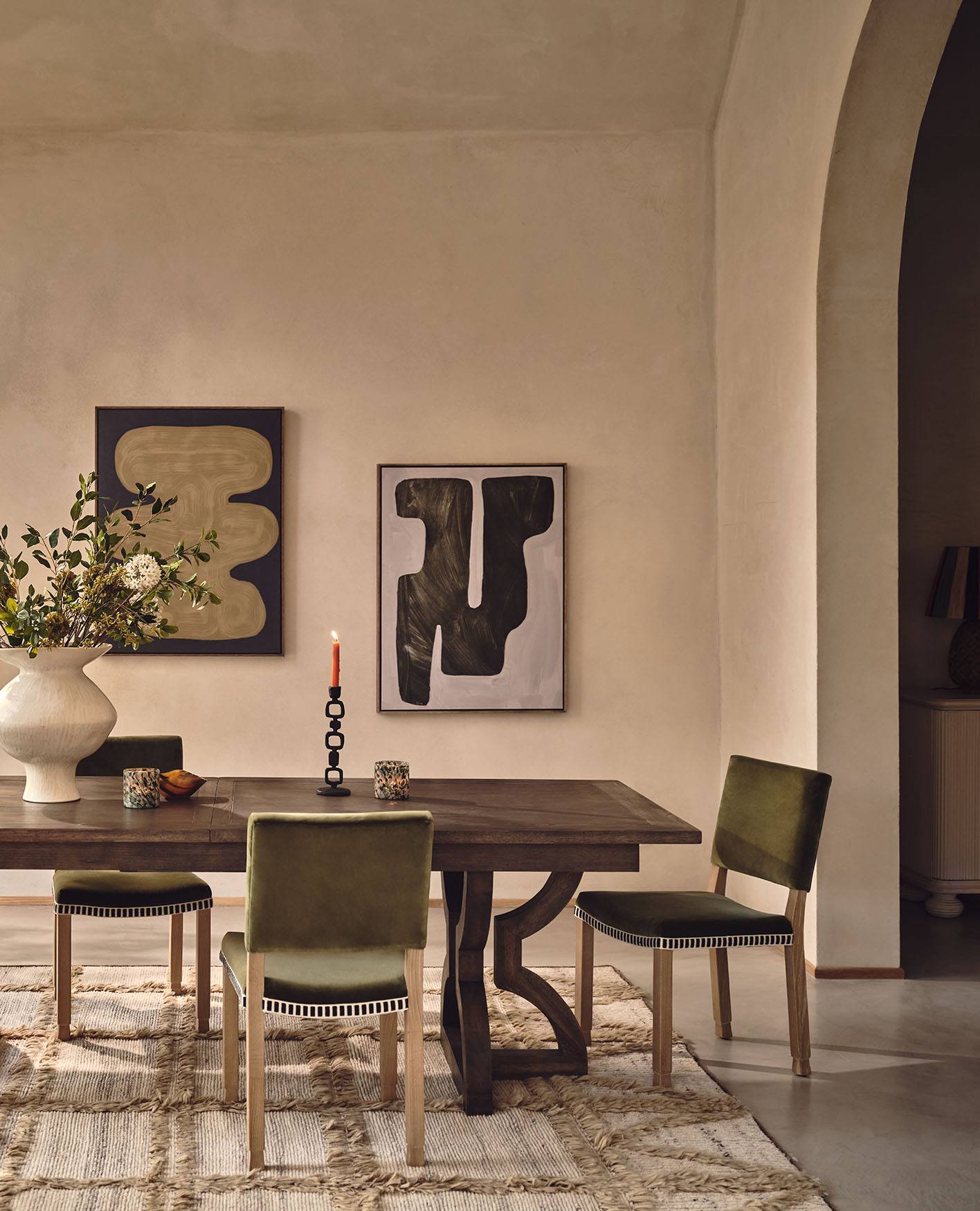 Green velvet dining chairs are arranged around a large wooden dining table. Graphic art prints decorate the plain plaster walls.