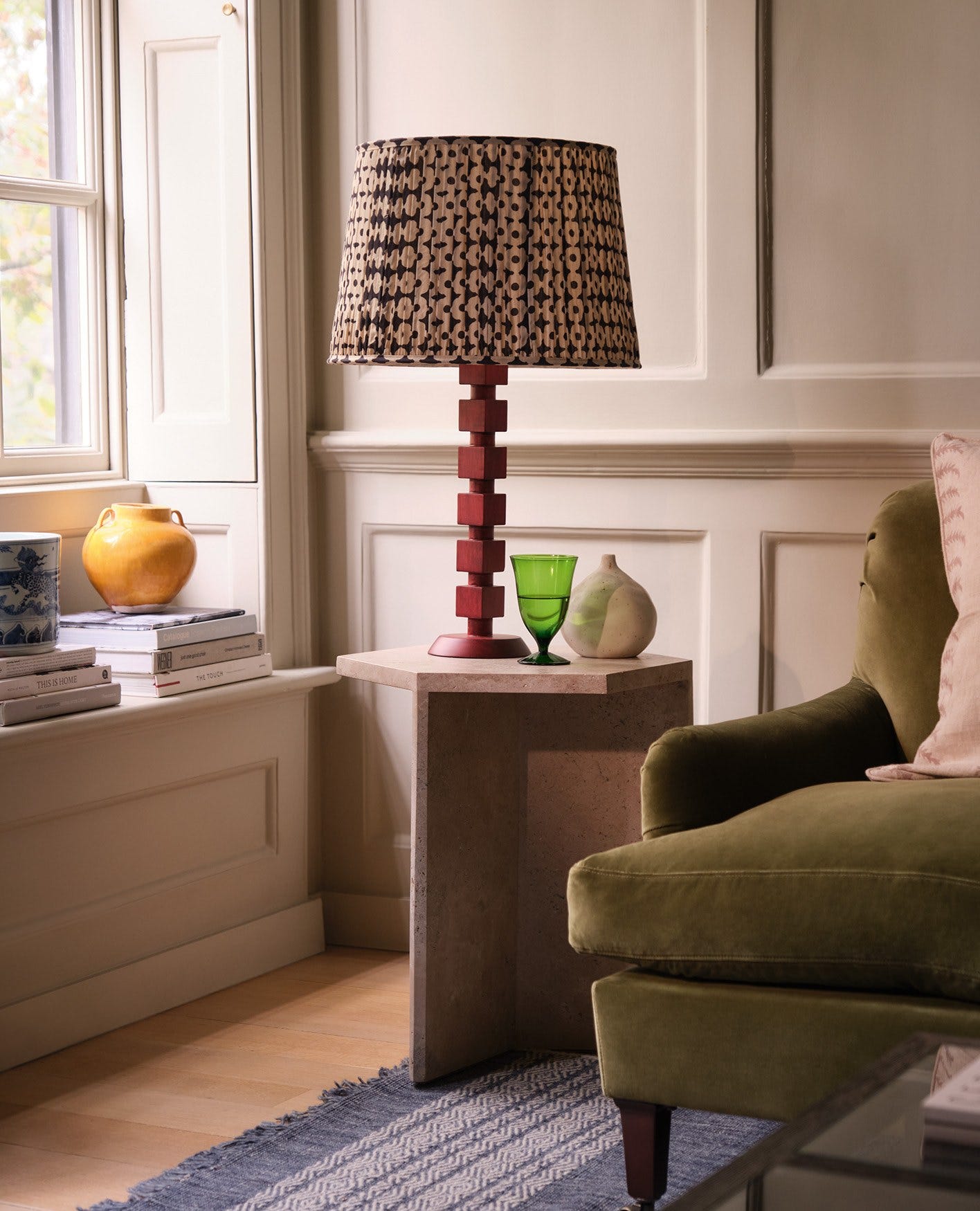 A red bobbin-inspired lamp with a pleated lampshade sits on a stone side table, next to a green velvet sofa.