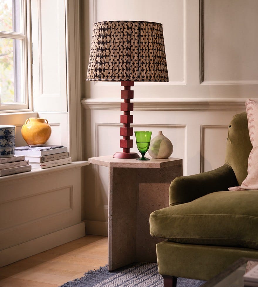 A red bobbin-inspired lamp with a pleated lampshade sits on a stone side table, next to a green velvet sofa.