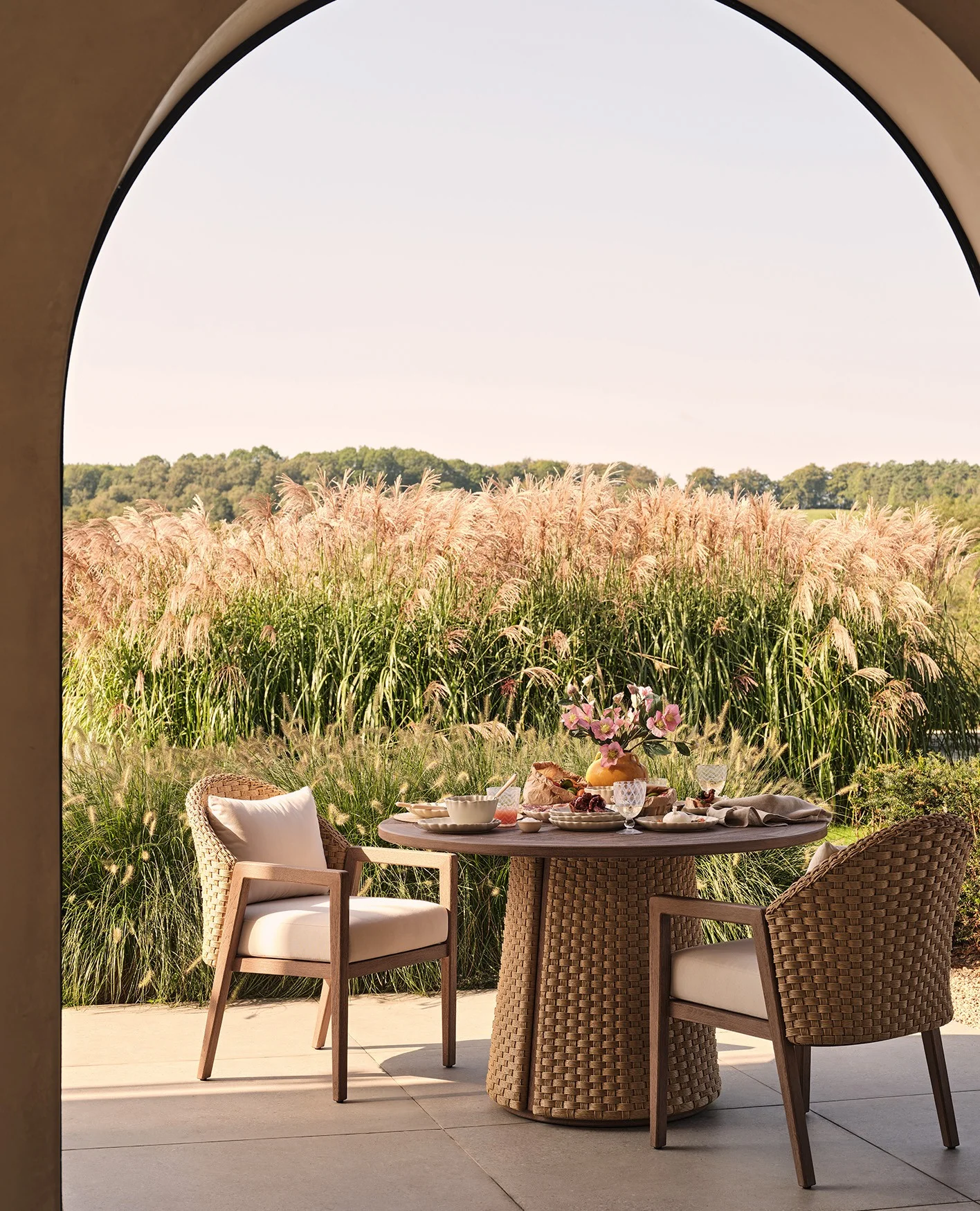 An outdoor dining set up with a round table and two chairs an be spied through an archway. Behind is a meadow and a blue sky.