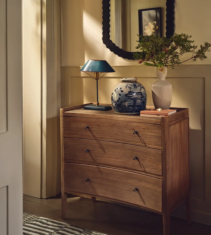 A chest of drawers sits in a hallway, decorated with a blue lamp and pot and a white vase. A black mirror hangs above it.