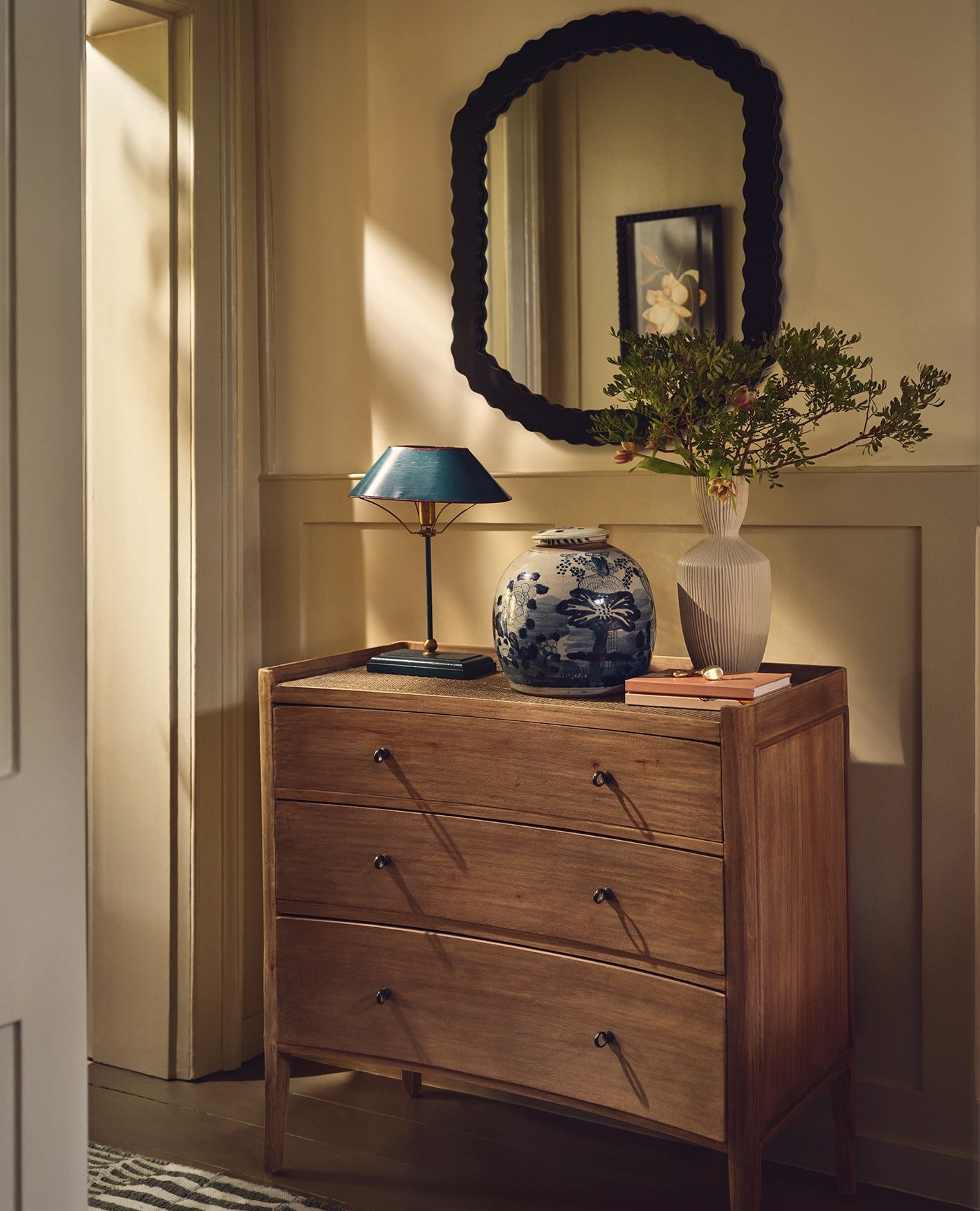 A chest of drawers sits in a hallway, decorated with a blue lamp and pot and a white vase. A black mirror hangs above it.