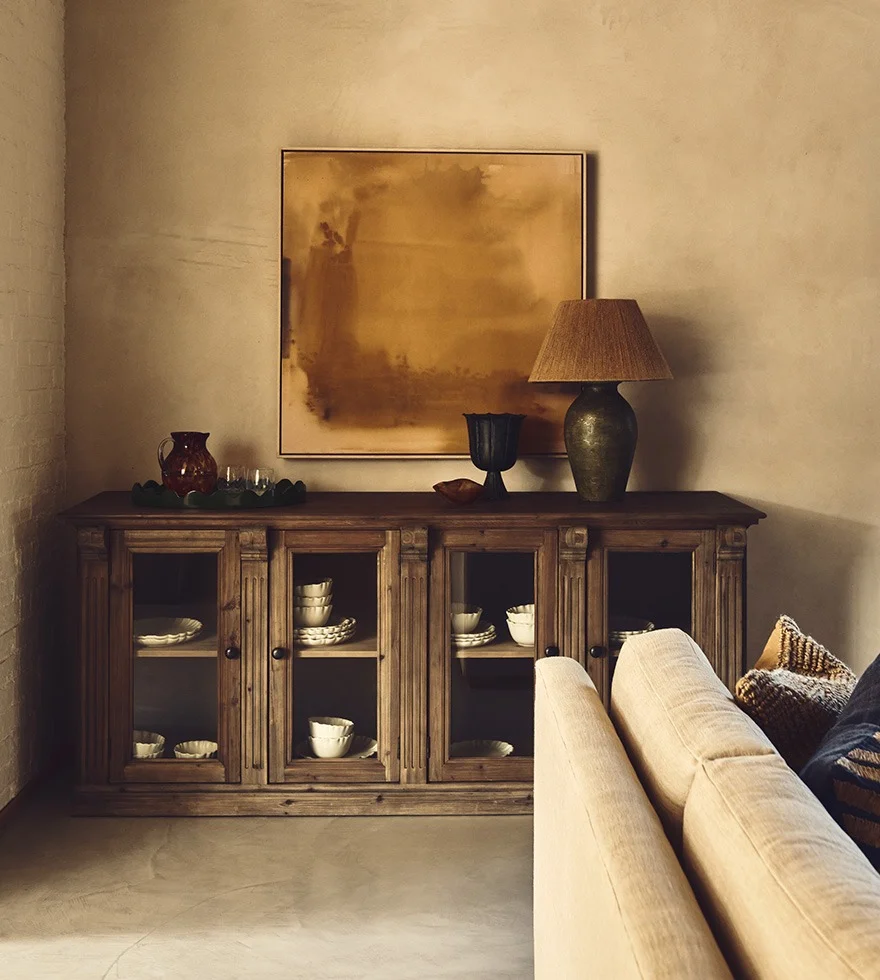 A wooden cabinet with glass-fronted doors sits in the corner of a sitting room. A mustard yellow painting hangs above it, and the corner of a white sofa can be seen in the foreground.