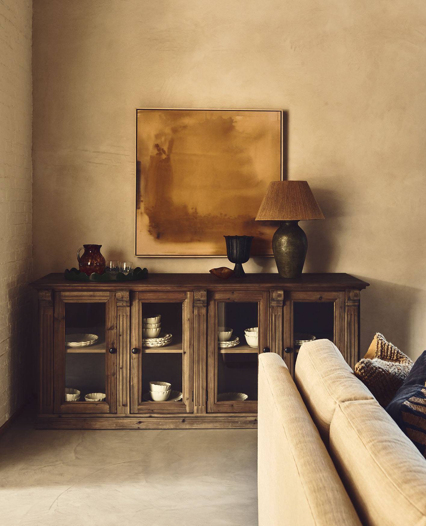 A wooden cabinet with glass-fronted doors sits in the corner of a sitting room. A mustard yellow painting hangs above it, and the corner of a white sofa can be seen in the foreground.