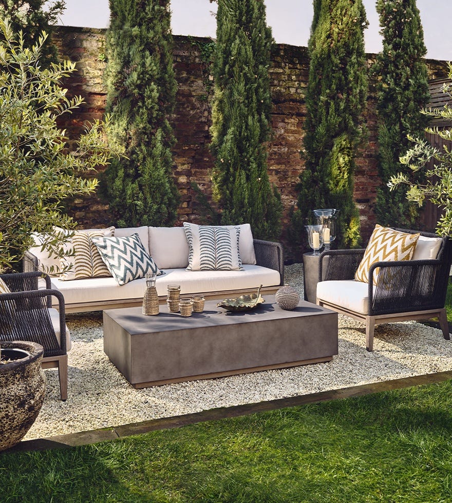 An outdoor sofa and outdoor armchairs surrounding a concrete grey coffee table in a leafy garden.
