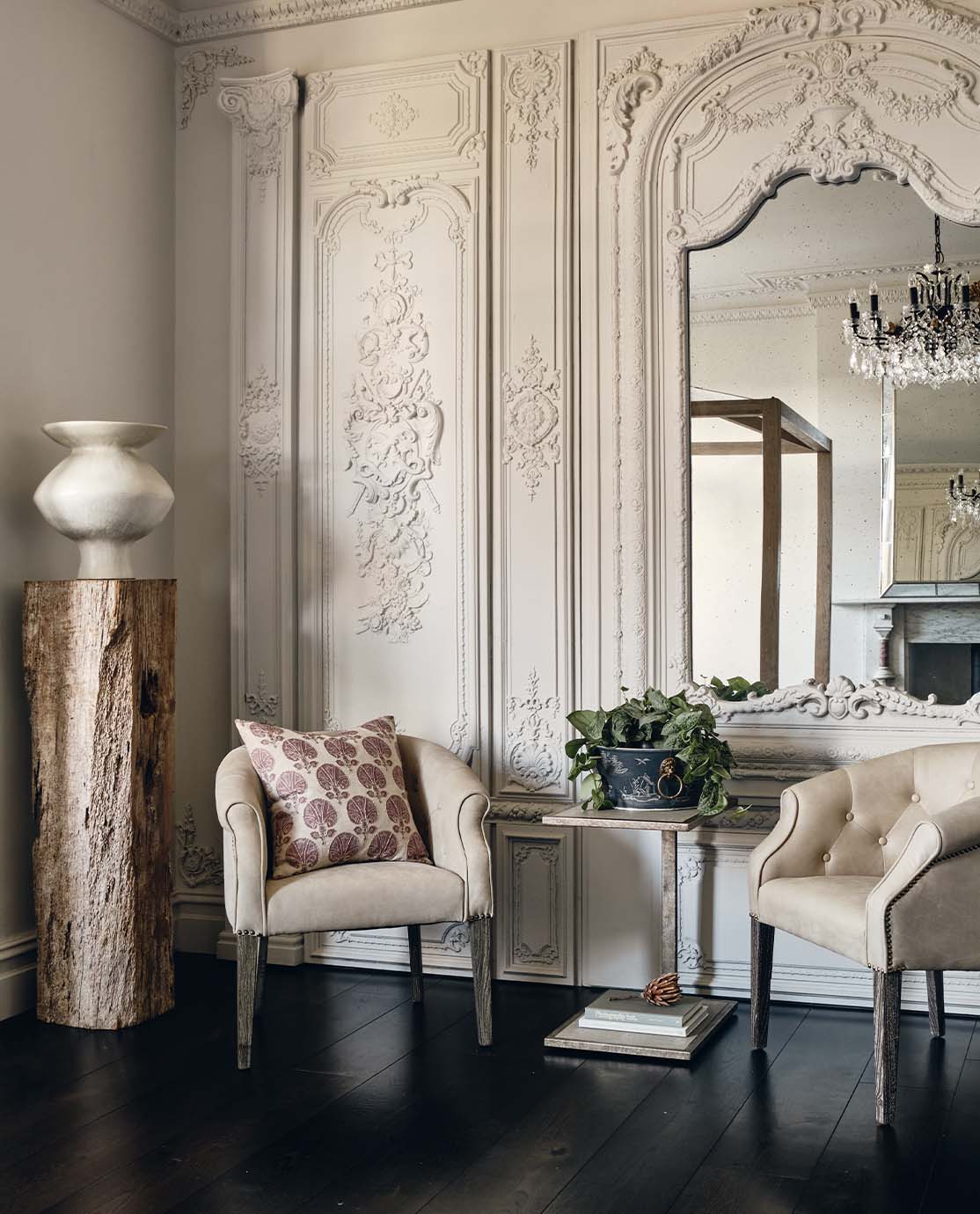 Two beige armchairs sit in front of an elegant wall, decorated with cornicing and a grand mirror