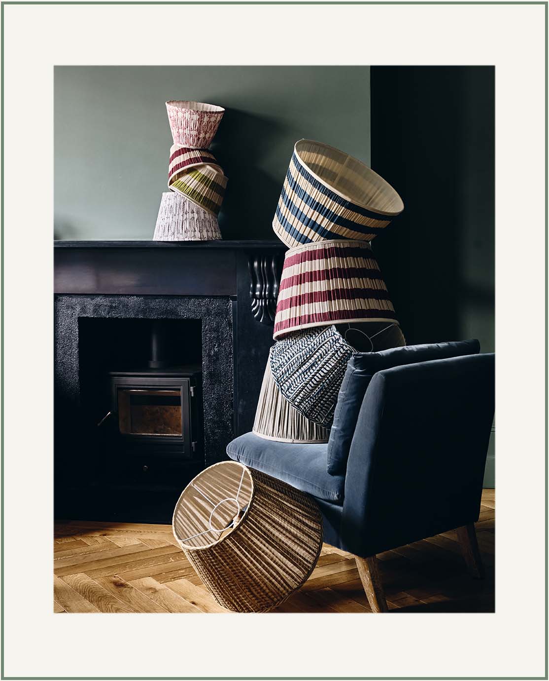 A tower of pleated and printed lampshades, artfully arranged on a velvet blue armchair and back mantelpiece