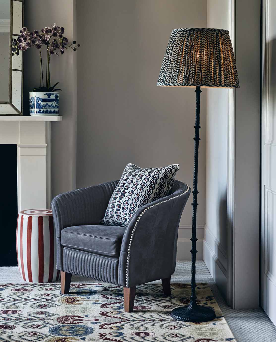 A skinny, black floor lamp decorated with a printed and pleated blue lampshade sits next to a blue leather armchair.