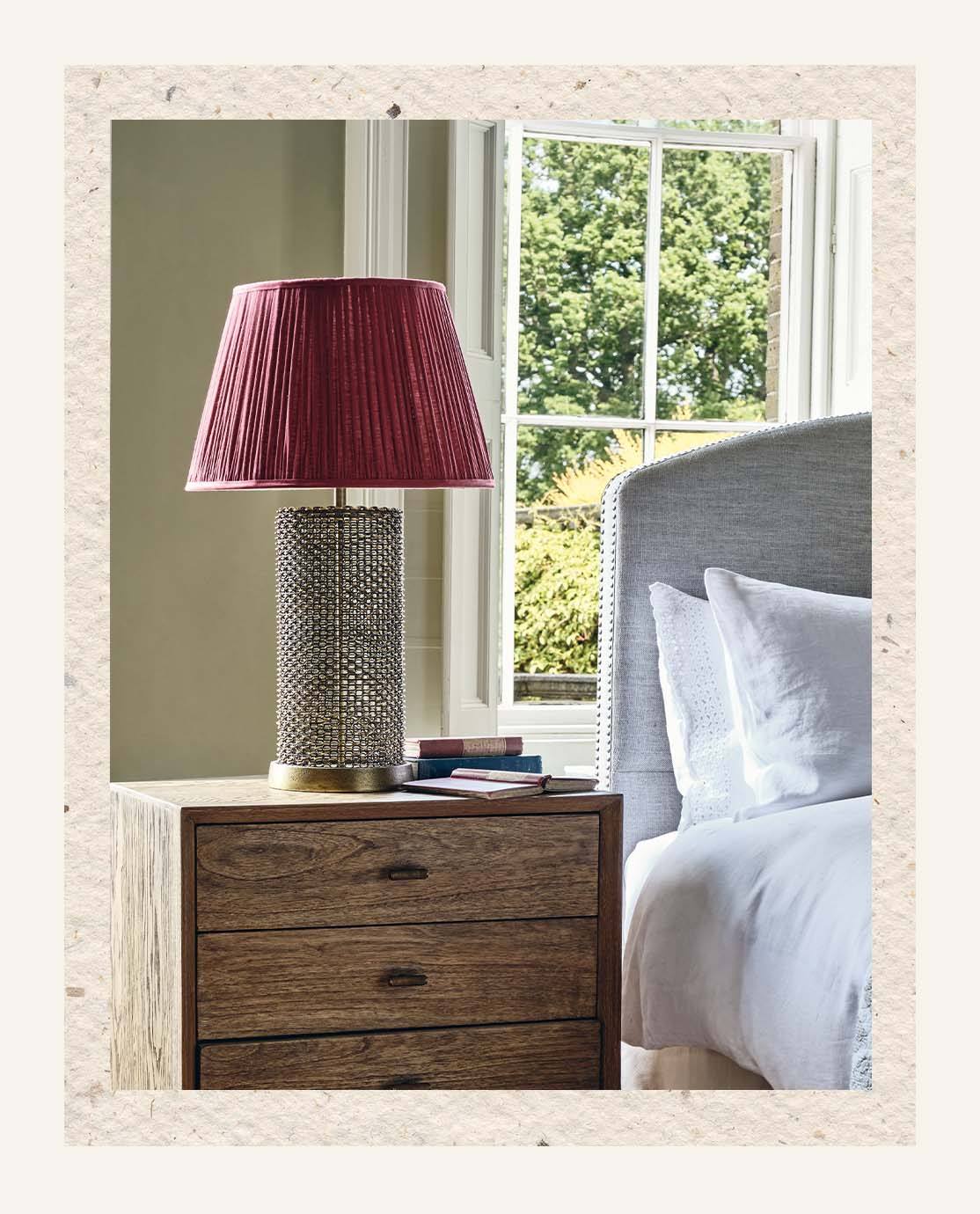 A sculptural lamp, decorated with a red pleated lampshade, sits on a wooden nightstand.