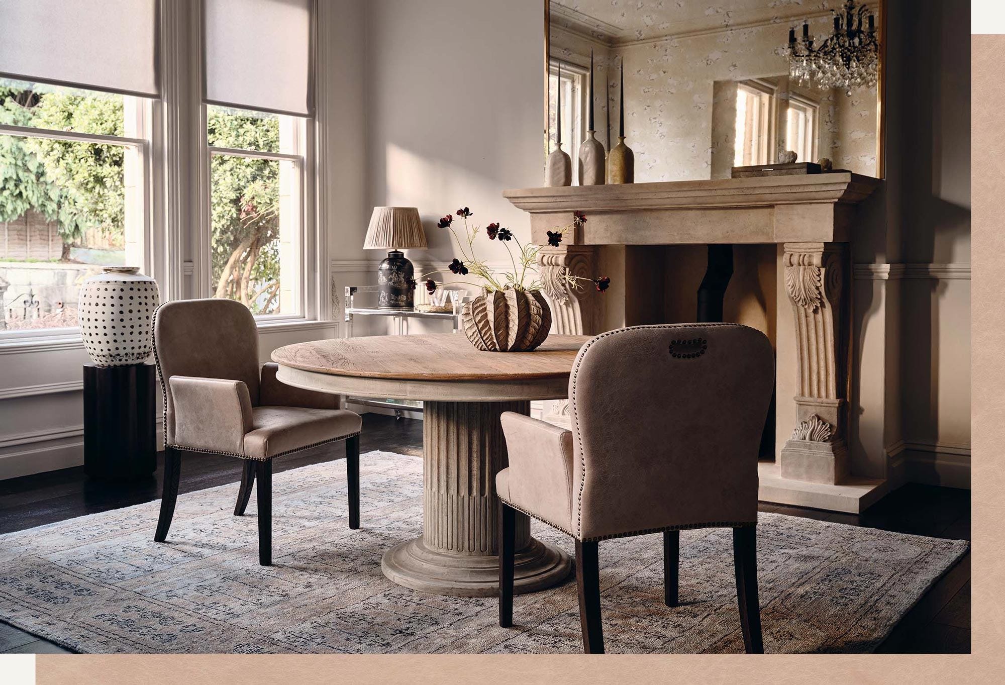 A dining room setting with a round table, beige leather dining chairs with arms and a mirrored bar cart in the background