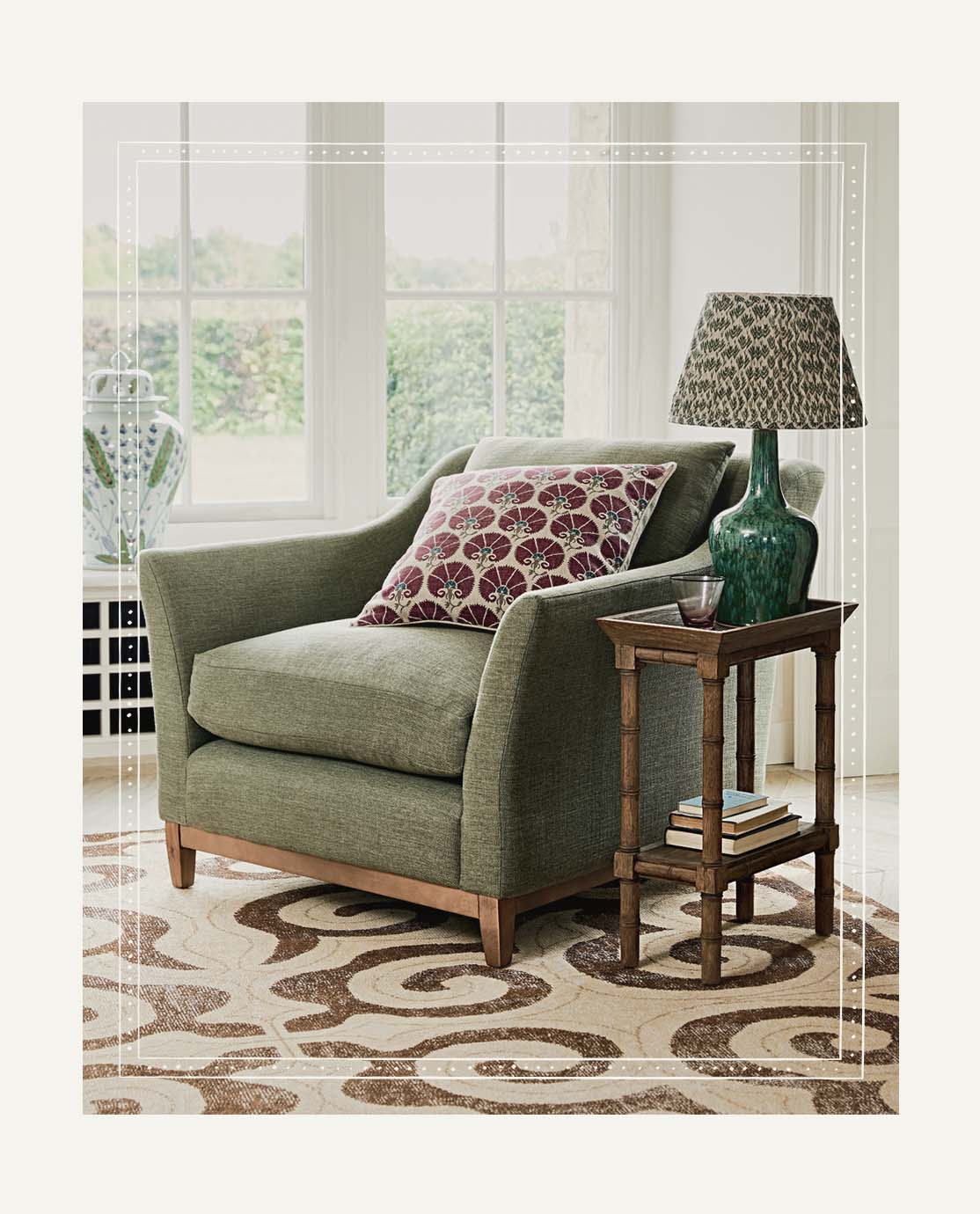 A green linen armchair with sloping arms sits on a patterned rug, next to a wooden side table and ceramic lamp.