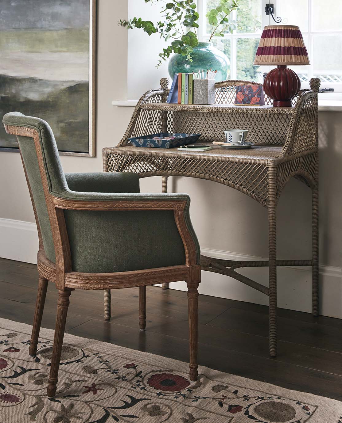 A green linen chair is pulled up to a rattan writing desk. A floral rug sits underneath it, and a landscape painting is hung to the side. 