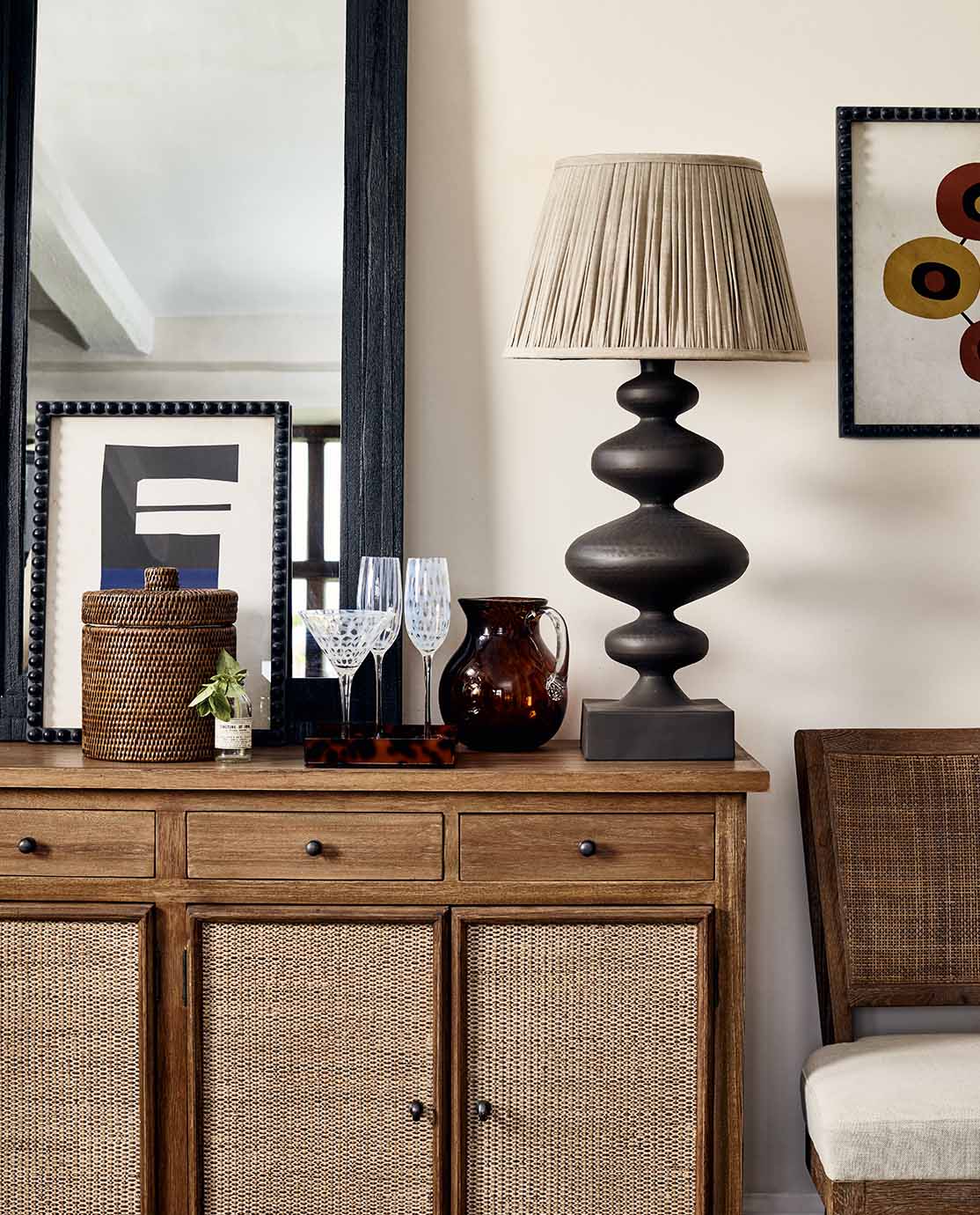 A rattan and wood console table is decorated with patterned glassware, a sculptural lamp and a rattan basket