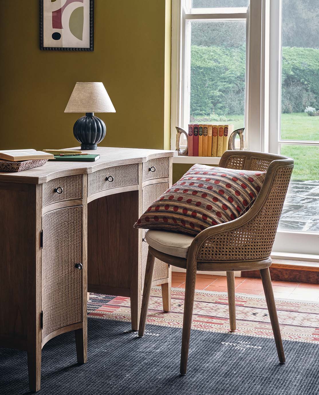 A cane dining chair decorated with a polka dot pillow is pulled up to a wooden desk, which holds a blue leather lamp.