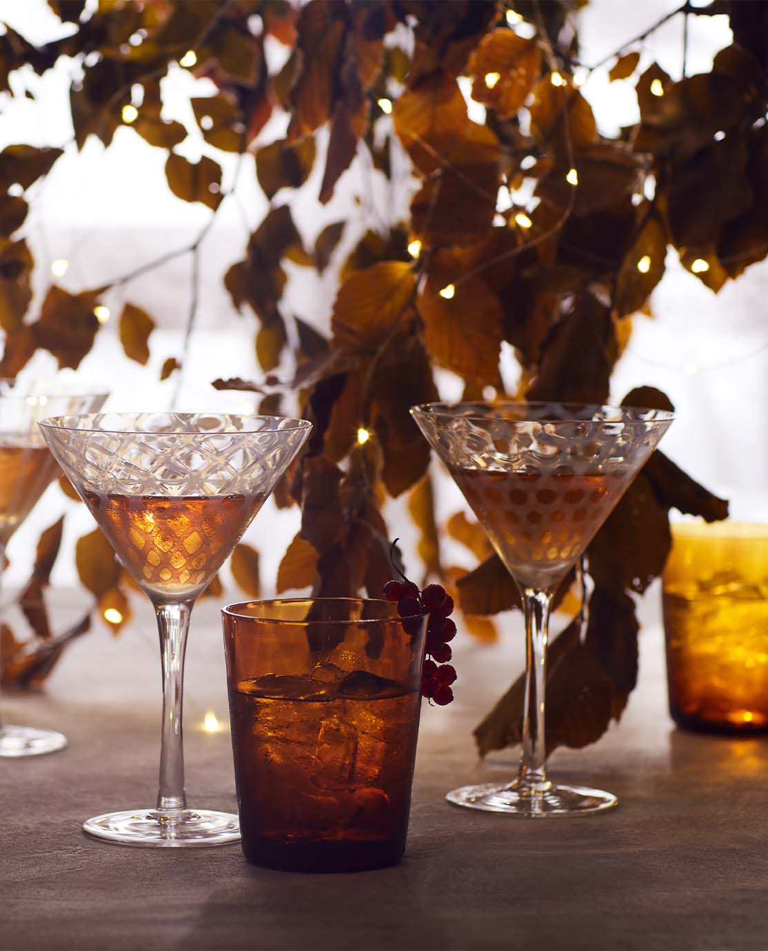 Two patterned cocktail glasses and an amber-colored tumbler sit in front of a branch of rust-colored leaves