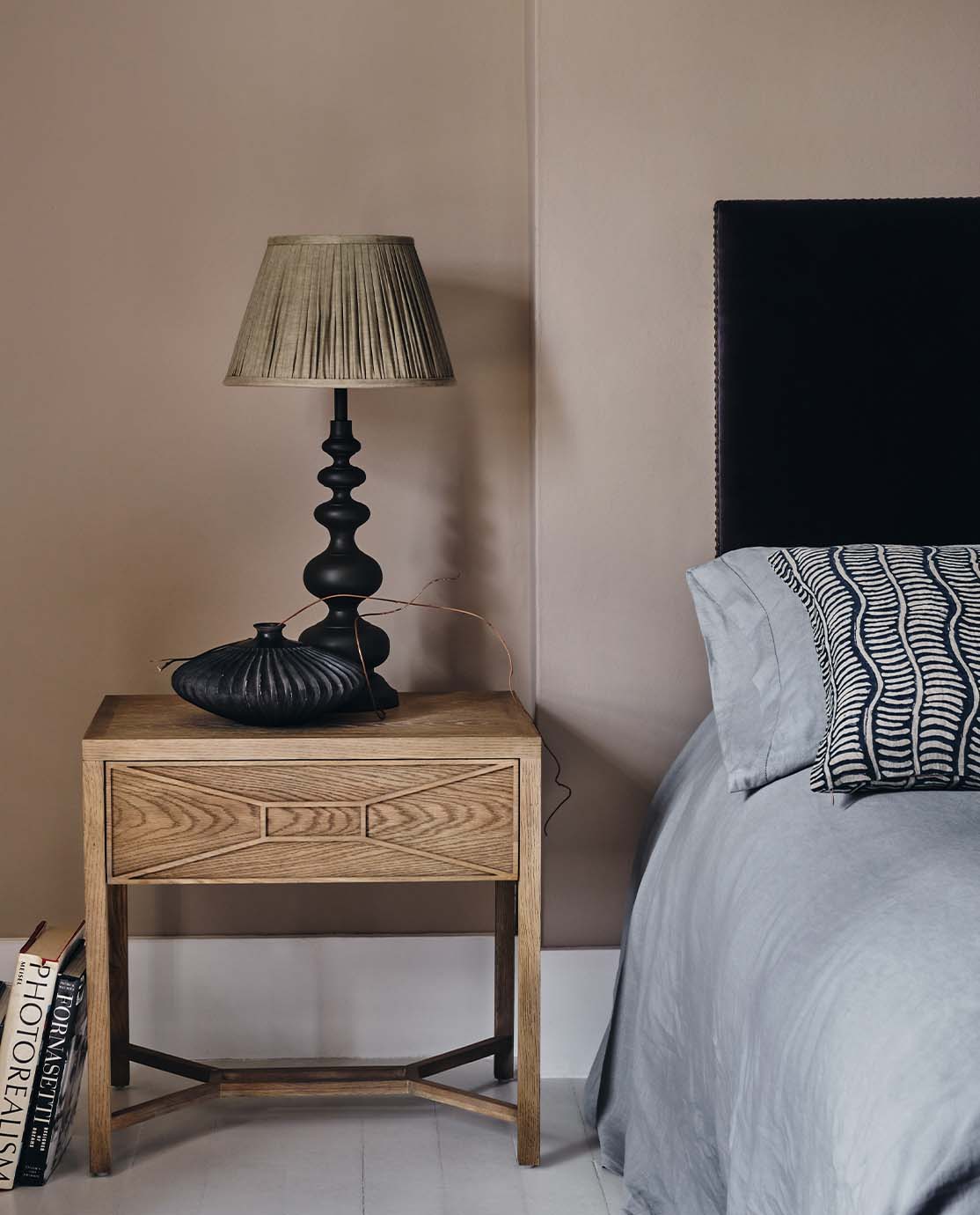 A black, sculptural lamp with a curvy base and grey pleated lampshade sits ona. wooden nightstand.