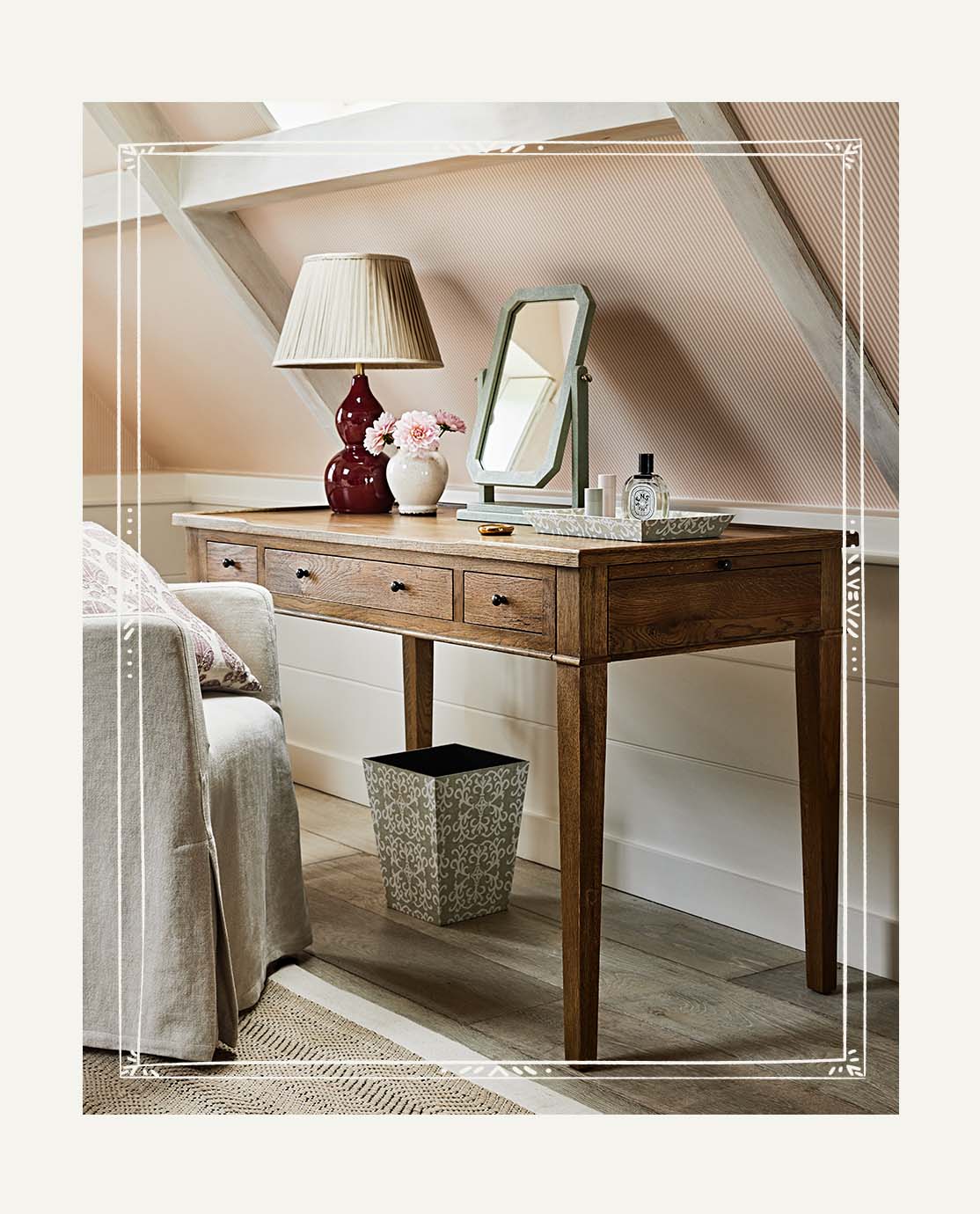 An armchair with a loose linen cover is drawn up to a wooden desk, which is decorated with a red lamp and faux shagreen mirror.