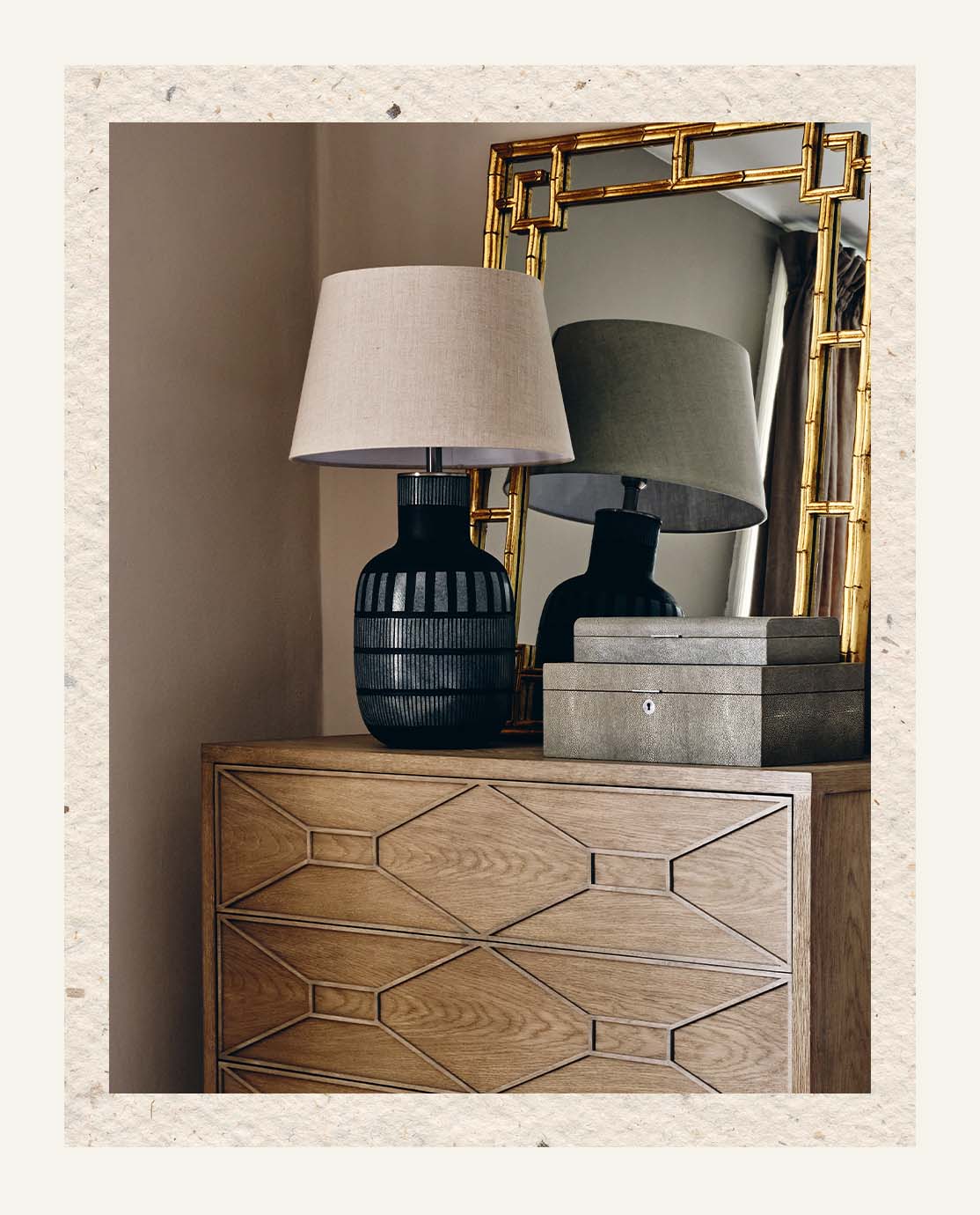 A black lamp with an abstract print sits on a wooden nightstand. A gold mirror sits behind it.