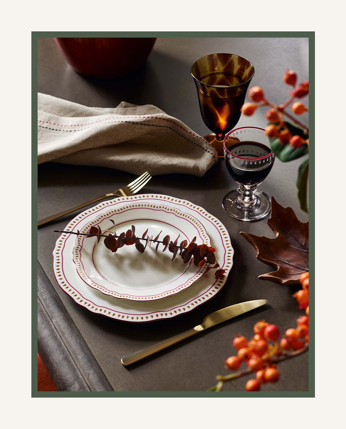 An autuman tablescape featuring white tableware, gold cutlery, red printed wine glasses and red berries, set on a black dining table