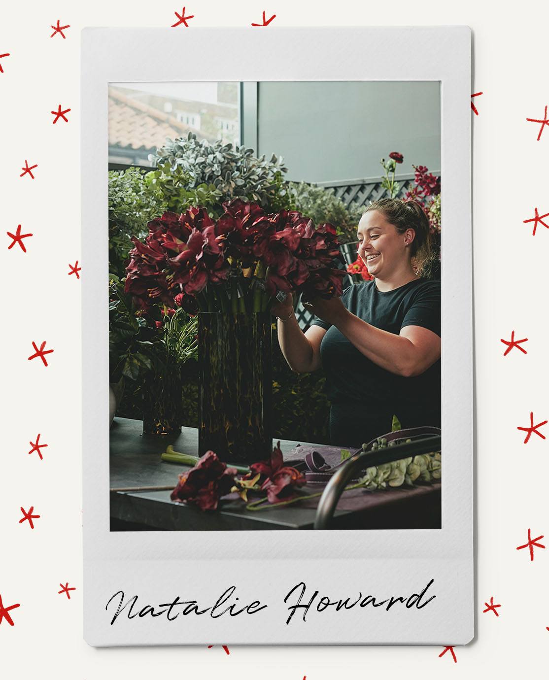 Natalie Howard wearing a black T-Shirt and smiling as she arranges from red and green flowers in a vase