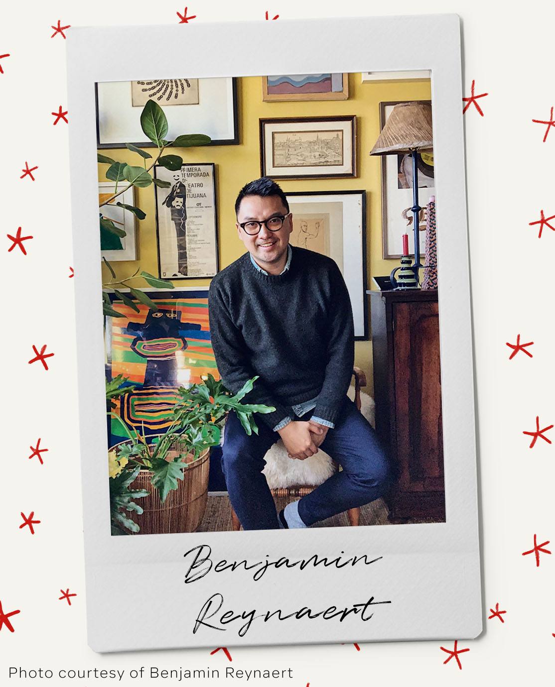 Benjamin Reynaert wears a grey jumper and blue shirt over dark blue jeans. He is sitting on a stool and smiling at the camera. Behind him is a yellow wall with a gallery wall of artwork.