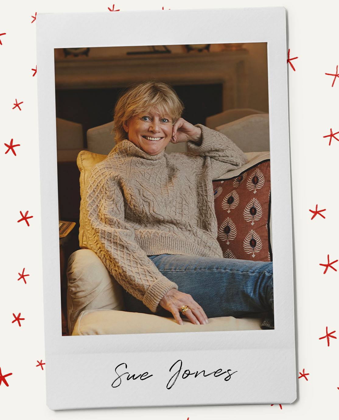 Sue Jones sits on a sofa wearing an aran knit polo-neck jumper and blue jeans. She is smiling at the camera and leaning on a rust-red cushion.