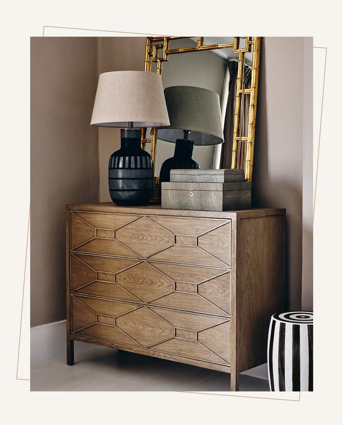A wooden chest of drawers decorated with diamond panel detailing. A navy patterned lamp and gold mirror sit on top.