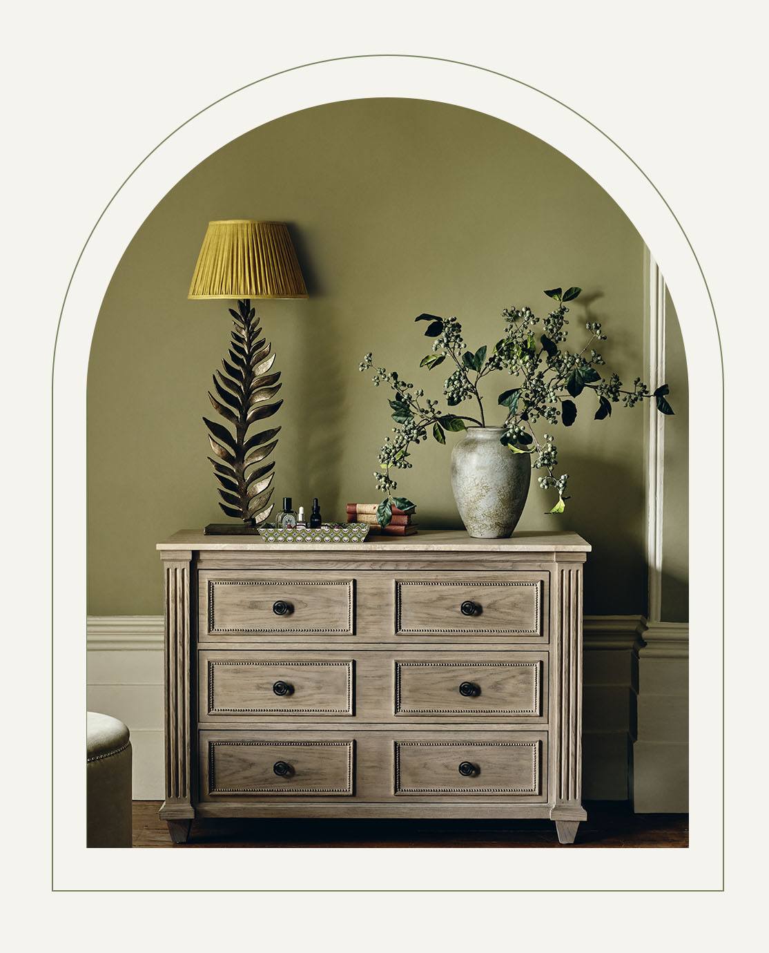 A blood orange velvet accent chair sits next to a wooden chest of drawers, which is decorated with a red lamp, a faux mini orchid and an artwork of fruit.