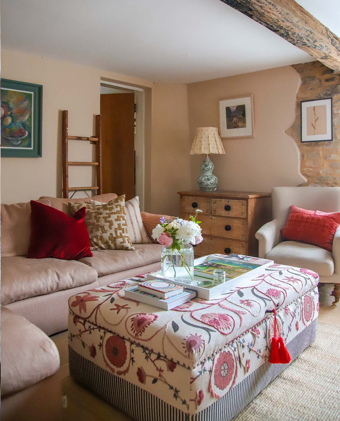 A cosy cottage sitting room with a pink L-shaped sofa, a red-and-pink embroidered ottoman and low wooden beams.