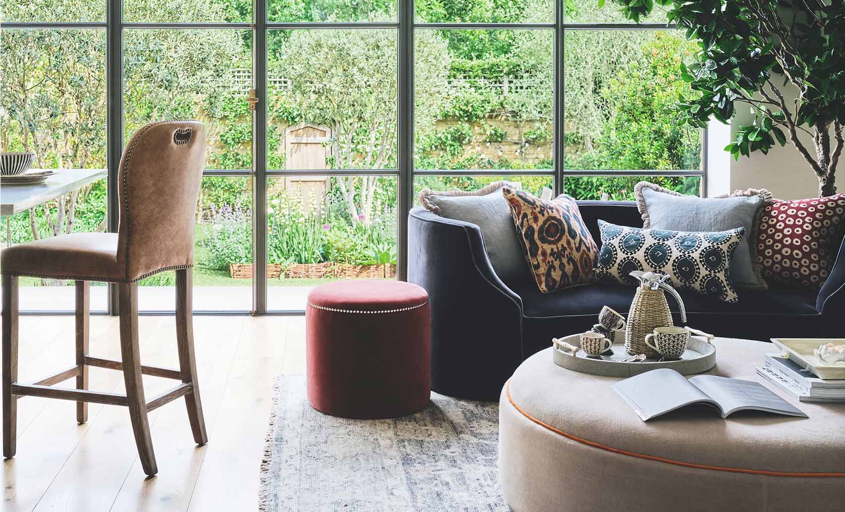 A velvet sofa decorate with lots of patterned cushions sits behind a circular fabric ottoman, which is decorated with books and crockery. There is also a red velvet ottoman in the photo and a cream leather bar stool.