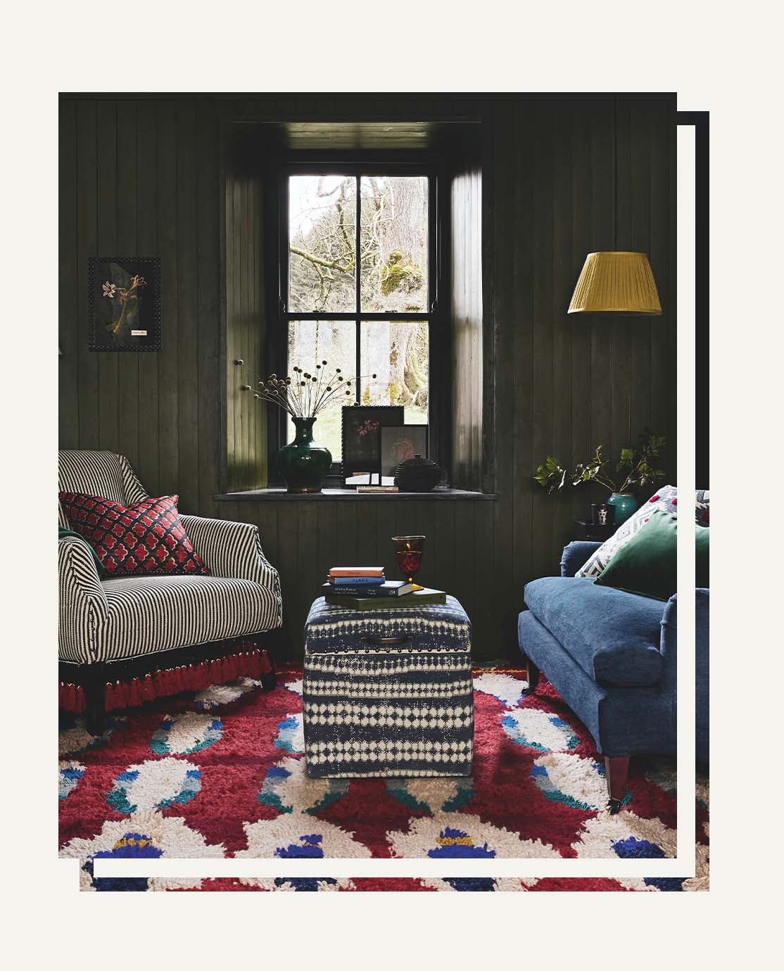 A sitting room setting with a navy blue sofa, a striped armchair with red tassels, a red patterned rug and a blue ottoman trunk with an abstract blue print