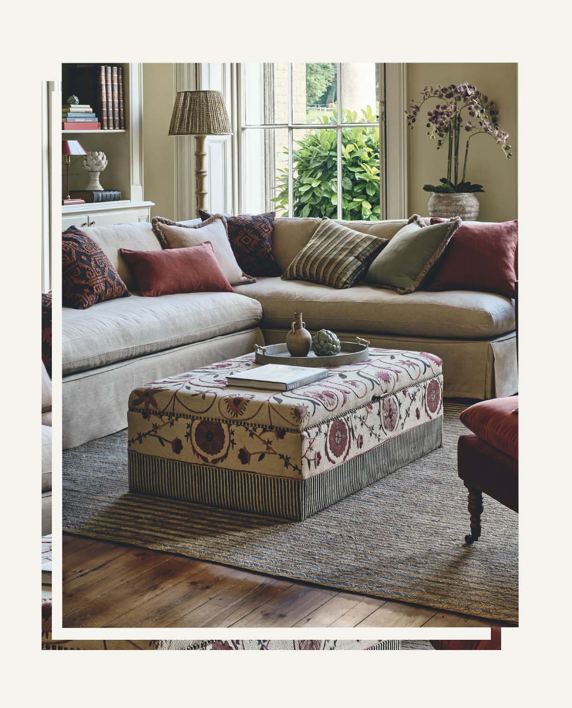 A beige ottoman with a red and green pattern sits in front of a beige sofa decorated with red and patterned pillows.