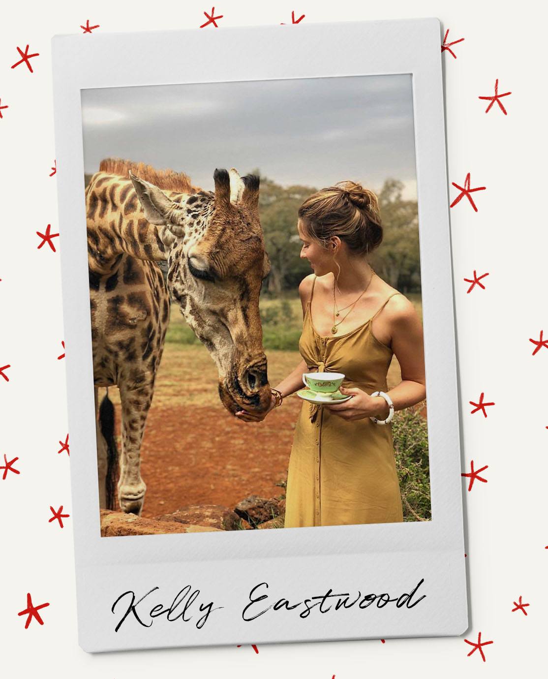 Kelly Eastwood wears a khaki silk slip dress. She is holding a blue china tea cup and saucer and looking away from the camera, at a giraffe that is directly behind her.