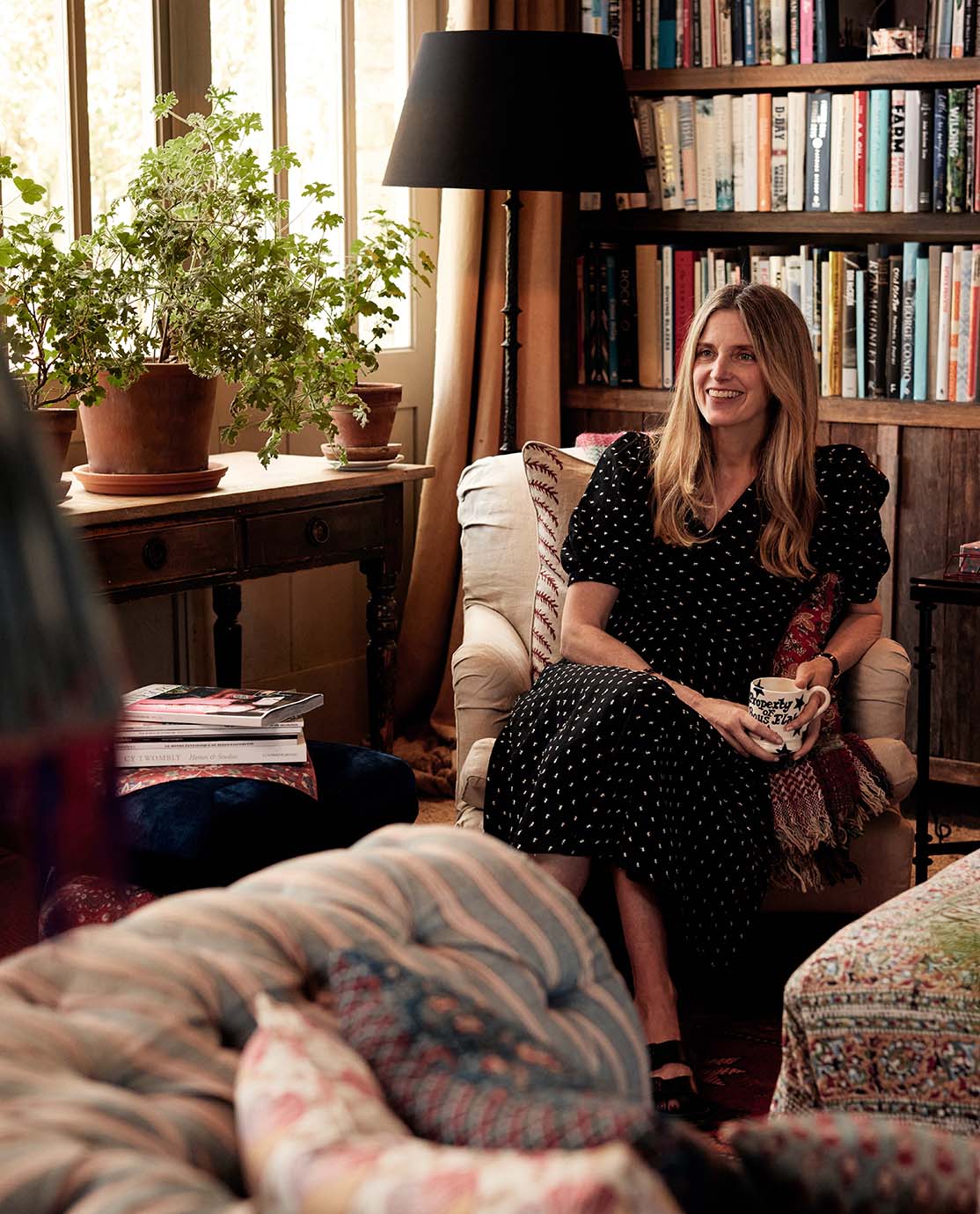 Amanda wears a black-and-white polka-dot dress. She is sitting in an armchair, smiling with a mug in her hand. 