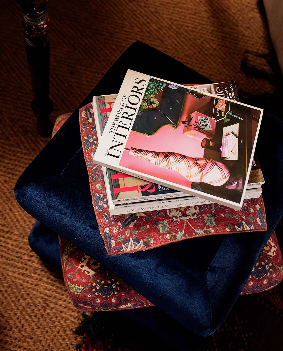 A stack of interiors magazines is placed on top of a red patterned pillow and a navy velvet stool.