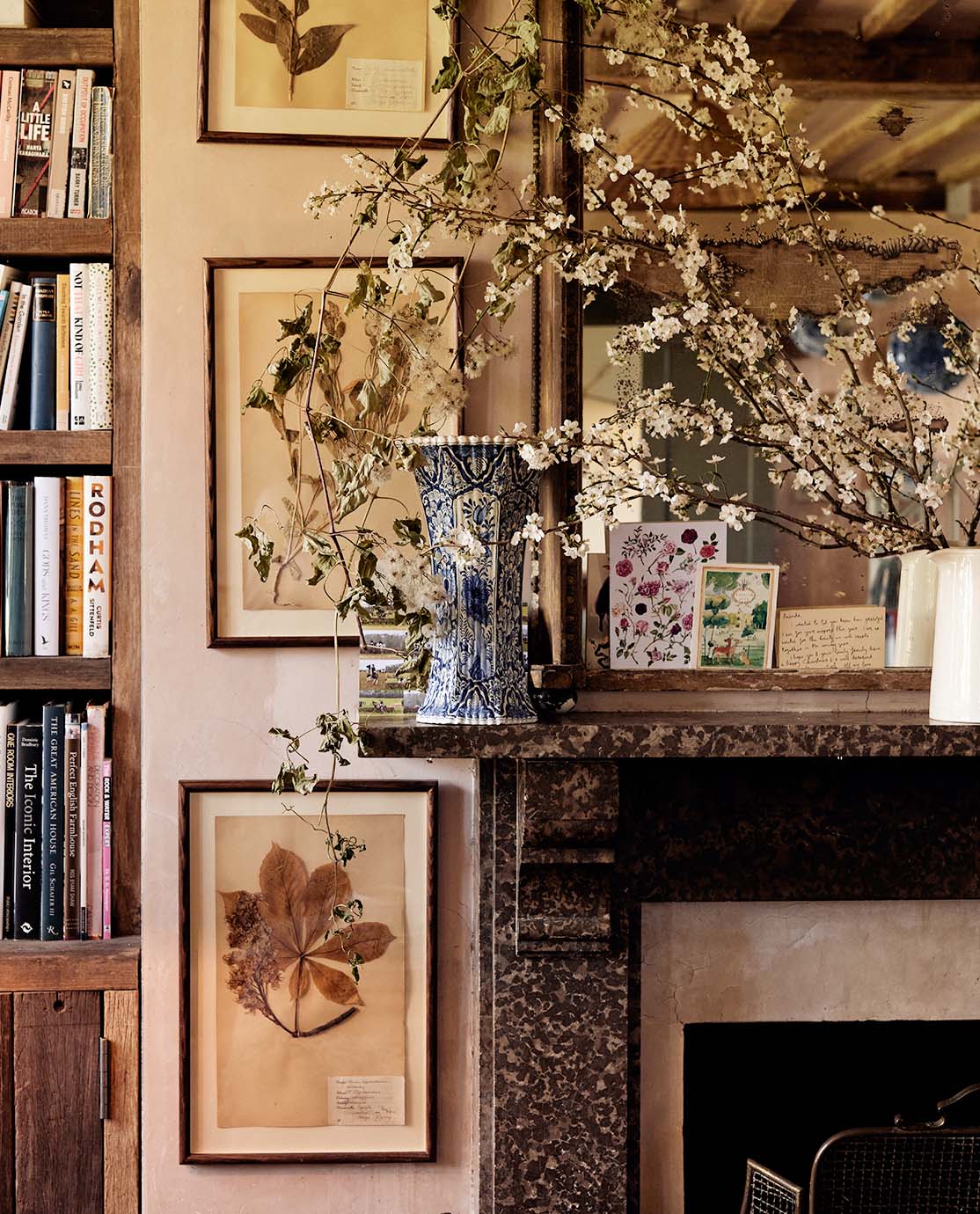 The corner of a mantelpiece decorated with a vase, a mirror and flowers. Three botanical prints hang on the wall next to it.