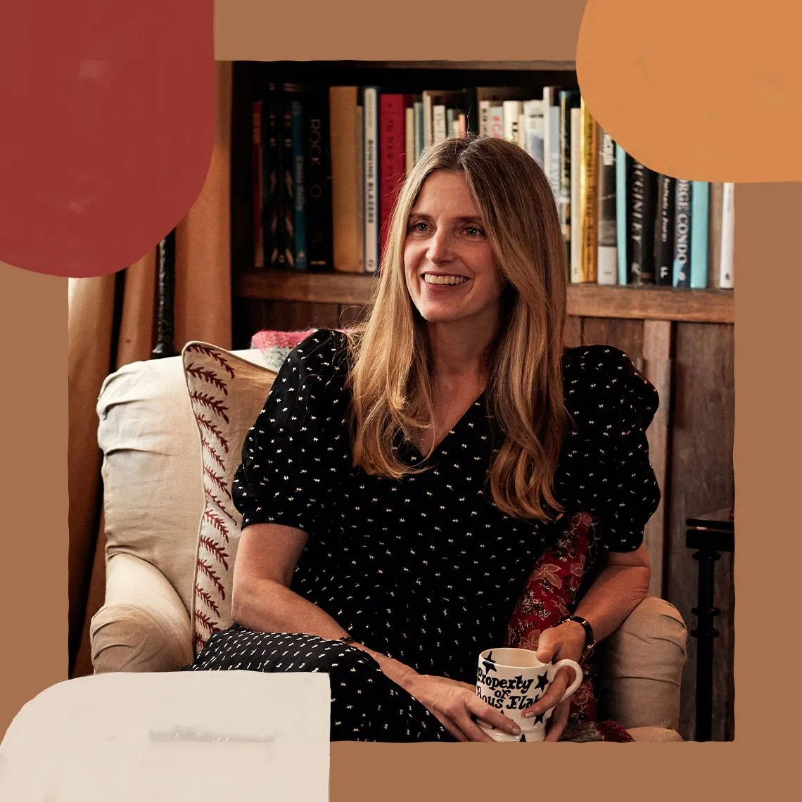 Amanda Cutter Brooks is wearing a polka-dot dress and is sitting in an armchair, holding a mug and smiling at somebody out of shot. A bookcase is in the background.